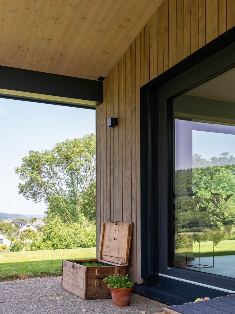 Holzfassade mit großem Fenster und überdachter Terrasse an einem Neubau in Holzbauweise