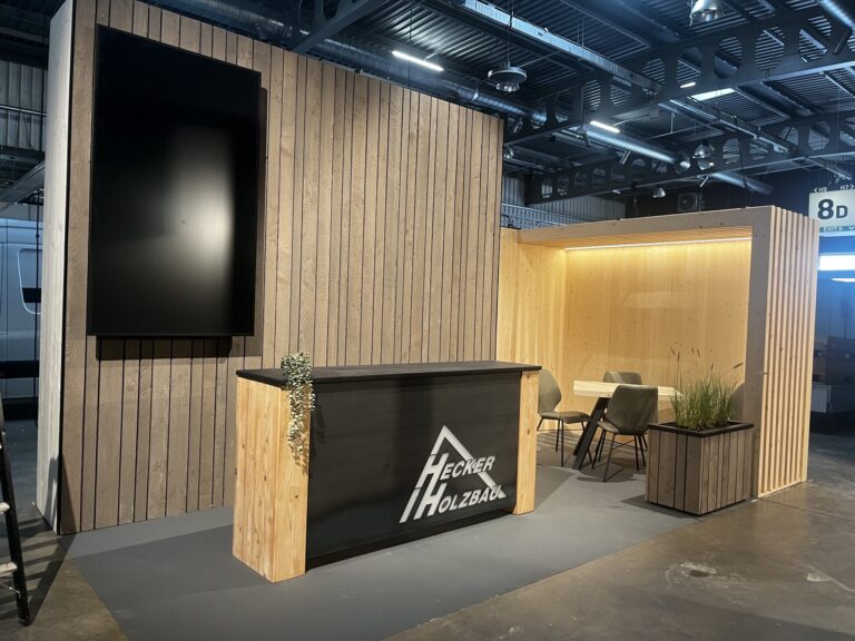 Messestand von Hecker Holzbau in der Messehalle: Holzwand mit Bildschirm, Theke mit Logo und Sitznische